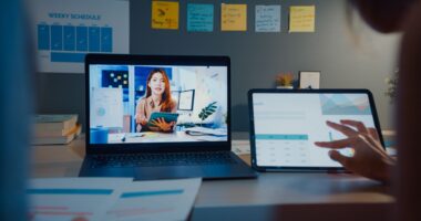 representação de um MBA online, em que uma pessoa assiste a aulas virtuais pelo computador e tablet. ao fundo, está uma parede repleta de post-its coloridos