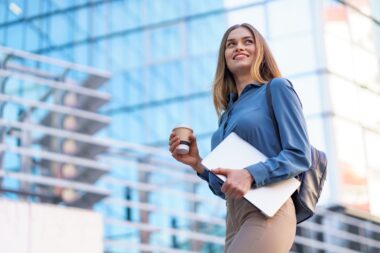 Mulher profissional sorridente com tablet e café, caminhando em frente a prédio corporativo, representando escolhas de carreira e as diferenças entre MBA e pós-graduação.