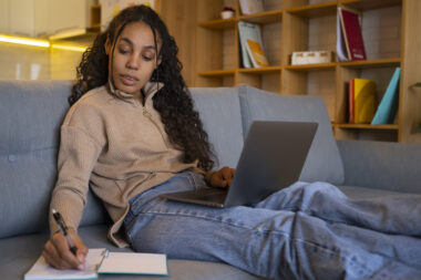 Estudar em casa: mulher sentada no sofá usando laptop e escrevendo em um caderno